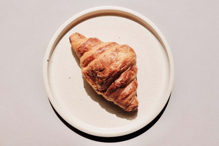 A kosher croissant displayed on a plate against a gray background. Richmond Kosher Bakery